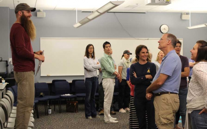 A person stands on a chair, speaking to a group of people at the family seminar of an outward bound intercept course